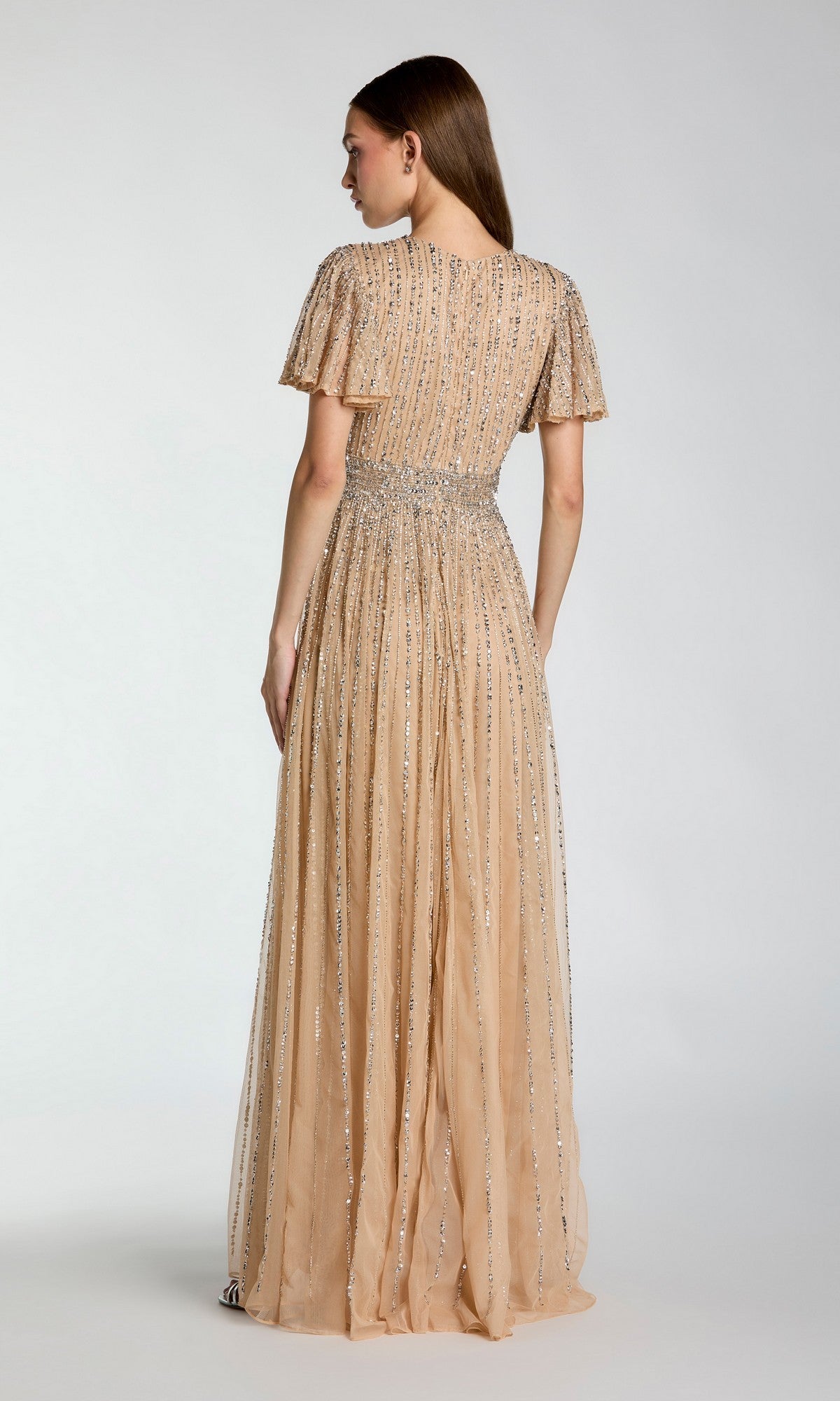 A woman is seen from the back, standing against a plain light background, wearing the Mac Duggal 5713 Long Formal Dress—an elegant peach, floor-length gown with short sleeves and vertical silver embellishments.